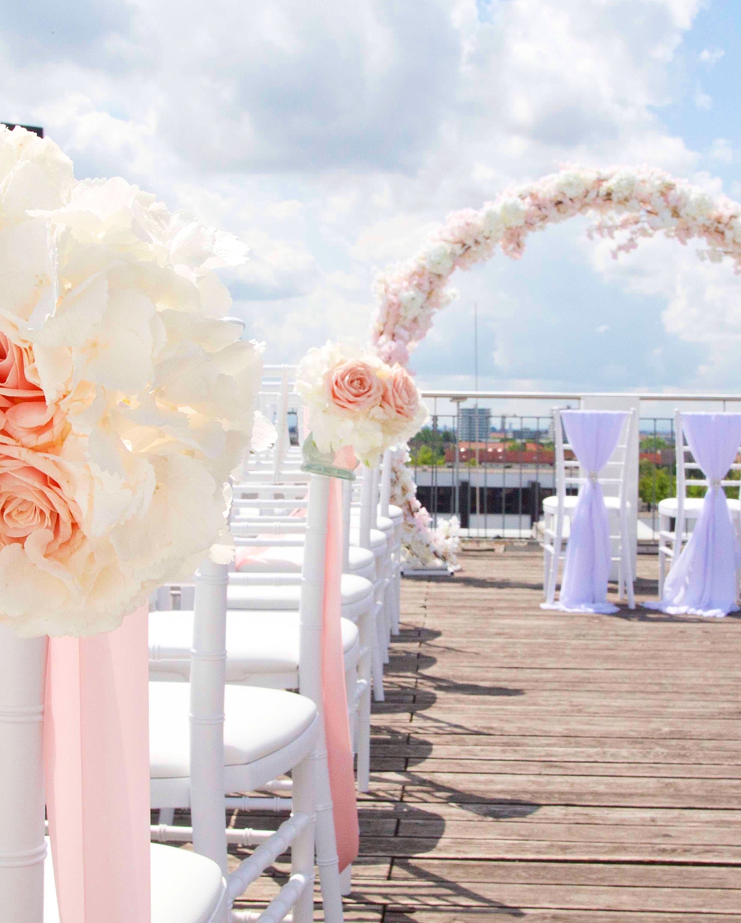 A rooftop wedding ceremony aisle: white chiavari chairs tied with blush and lilac ribbon and topped with cream hydrangea and peach rose posies, leading to a floral arch under a bright cloudy sky